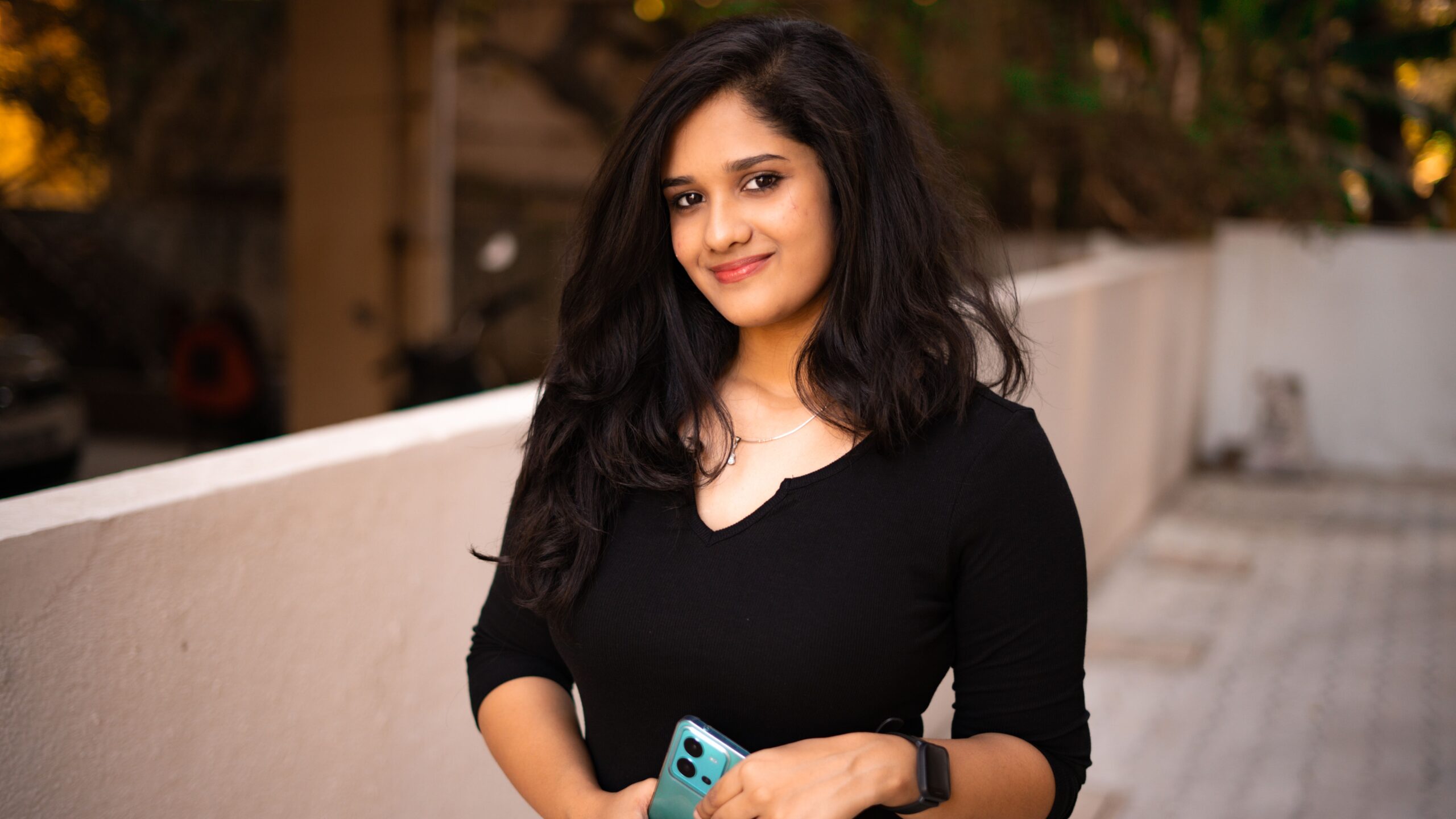 A young woman with long hair smiles while holding a smartphone outdoors.